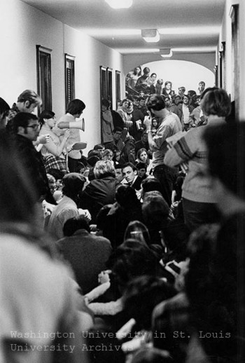 A photo of the 1968 student takeover of Brookings Hall. Students are shown crammed into the hallway, many sitting, with one student using a loudspeaker. 