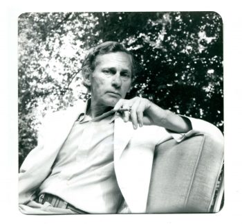 An image of William Gaddis from 1990 where he is lounging outside with a cigarette in-hand. 
