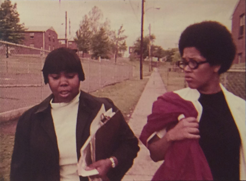 Two African-American women walking down a sidewalk. Both women are smartly dressed and one carries a clipboard filled with papers. 