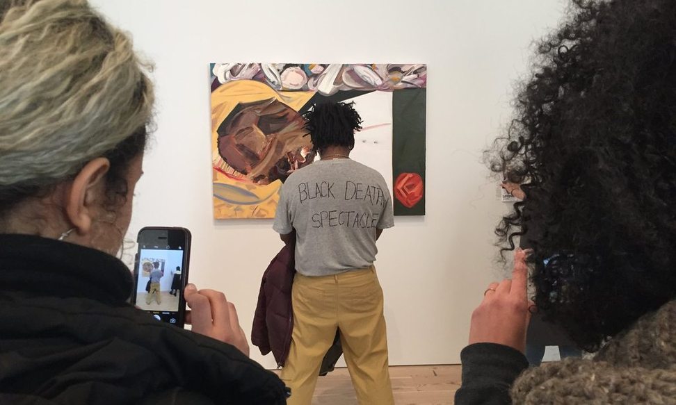 Scott Young stands in front of Dana Schultz' painting of Emmett Till, Young's back facing the photographer. The back of Young's shirt reads "Black Death Spectacle" in protest of the painting. 