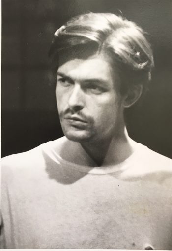 Headshot of the actor Lee Kissman during a scene in My Daddy is Dying. Kissman has a serious expression on his face as he looks into the distance; he is wearing a white t-shirt and sports a small mustache for the role. 
