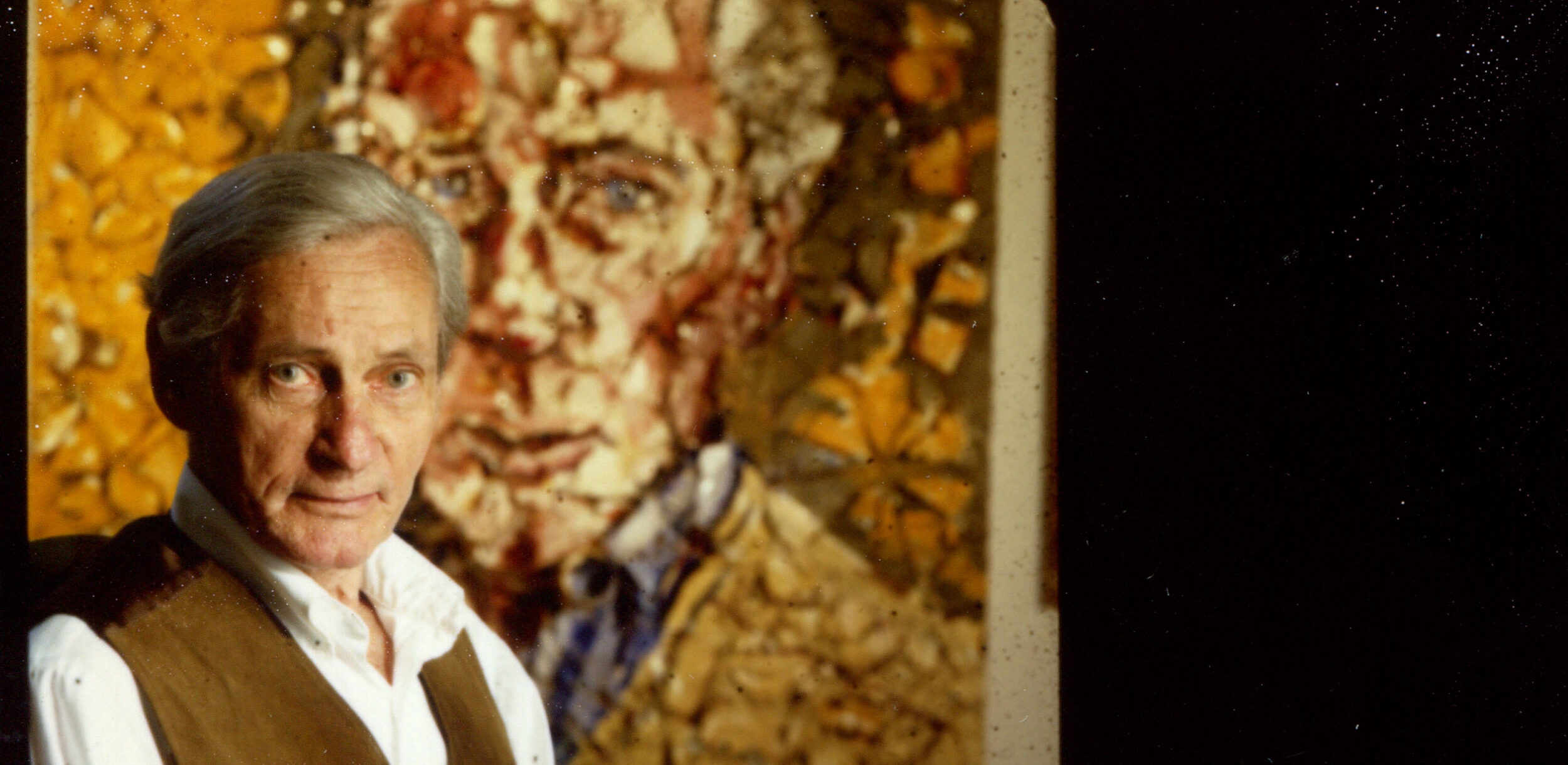 A photo of William Gaddis seated in front of a portrait of the author.