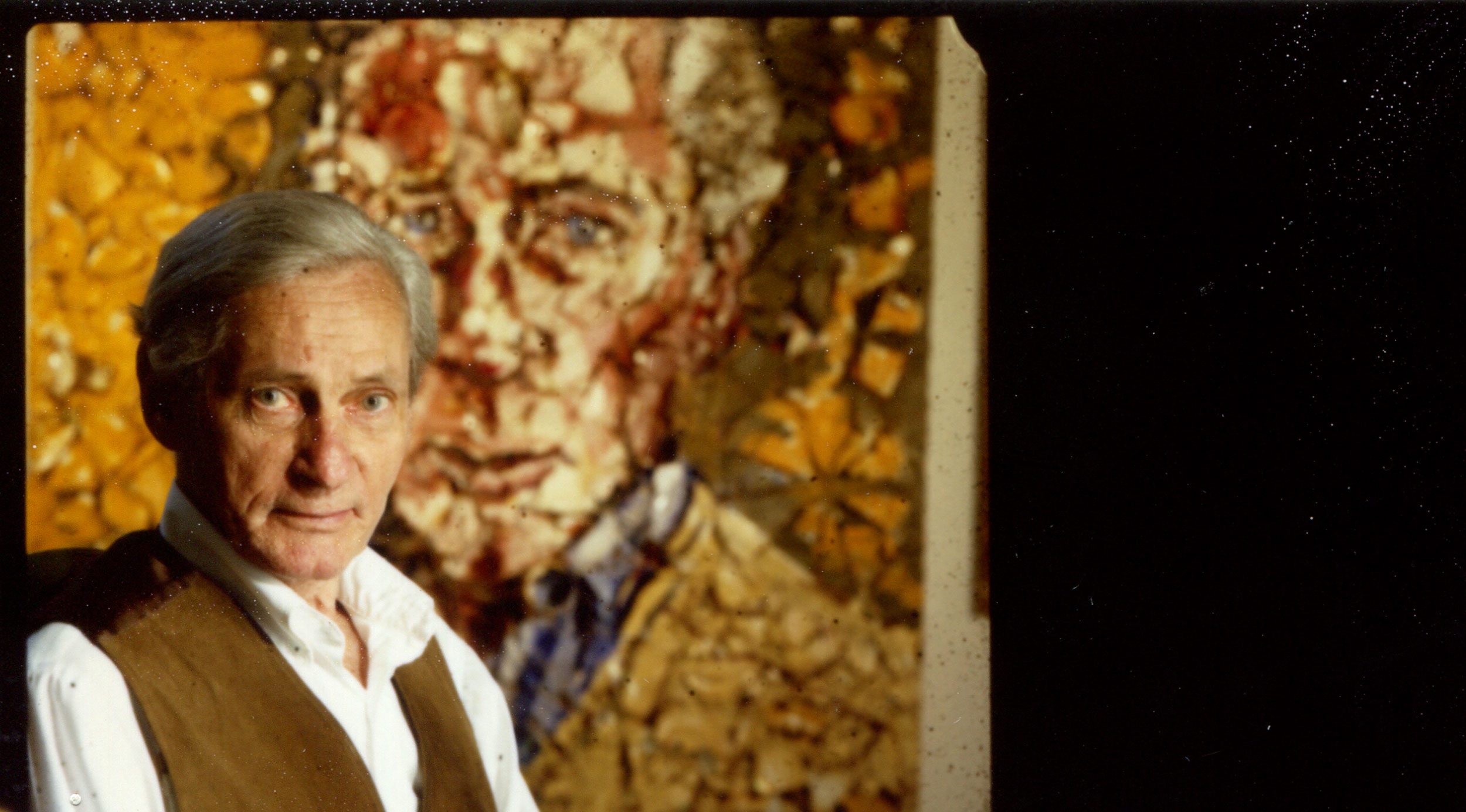 A photo of William Gaddis seated in front of a portrait of the author.