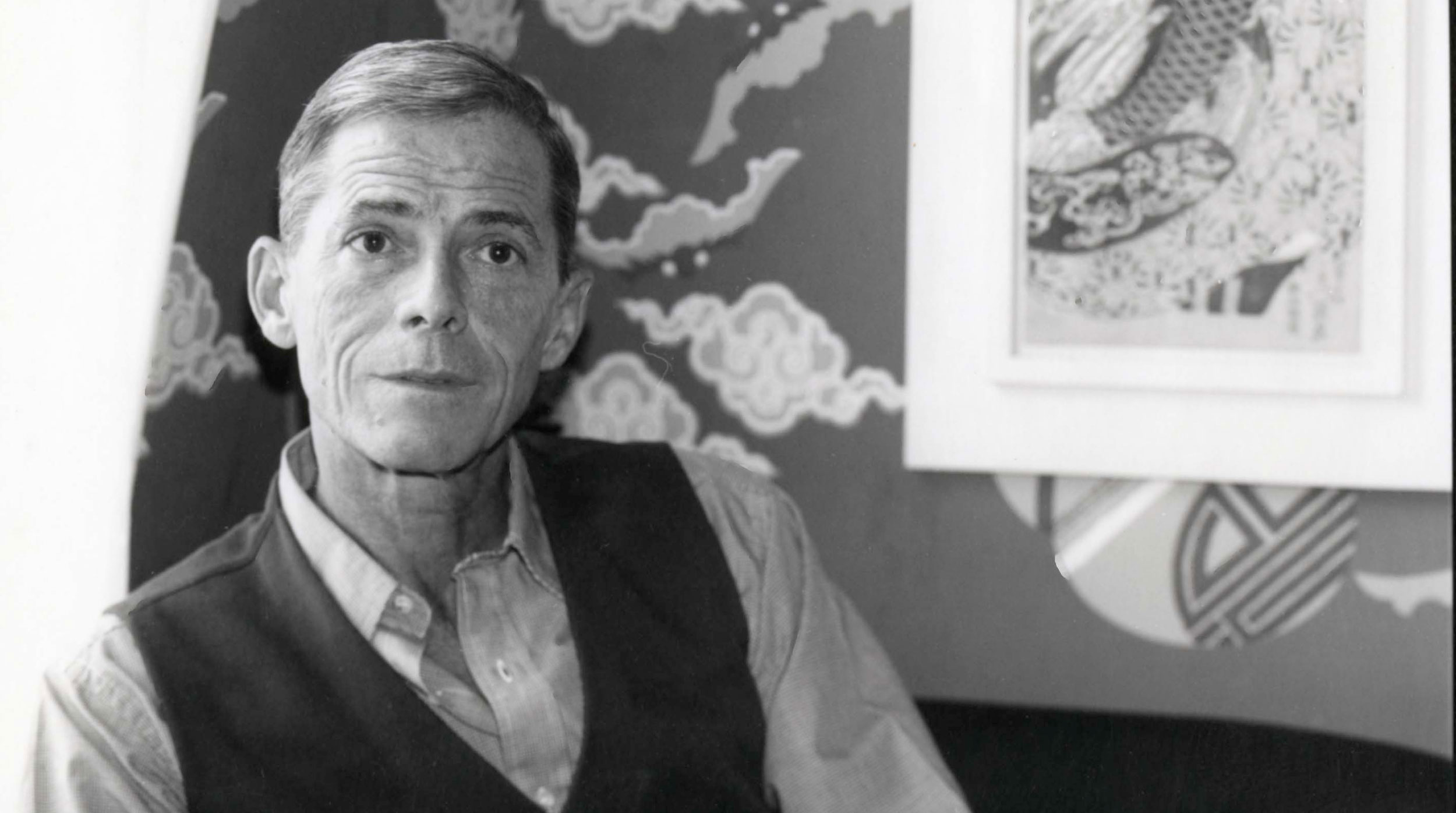 James Merrill sitting in a button-up beneath a vest in a wallpapered room.