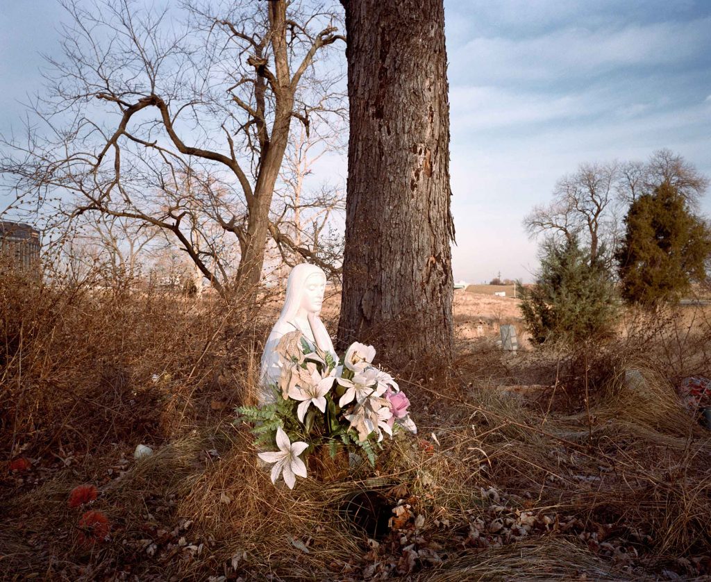 A grave site with a mother Mary statue standing as a marker. The grave has fresh flowers, but is otherwise in an overgrown location.