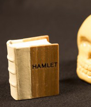 A small, miniature book of Hamlet alongside a small, wood carving of a skull. 