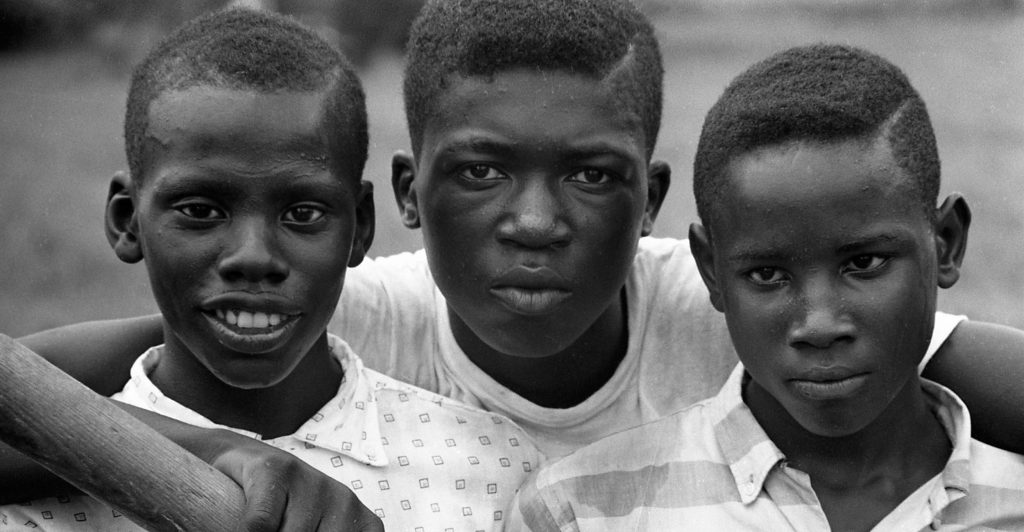 Three young Black boys standing with the arms of the middle figure over the shoulders of the other two as they look at the photographer. 