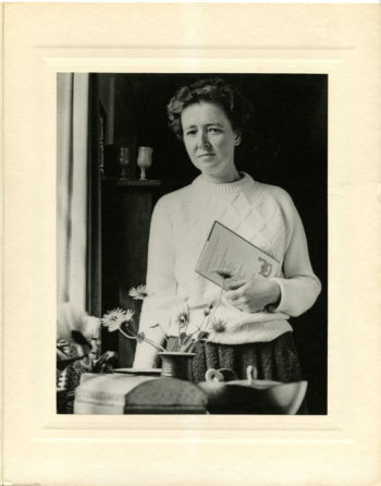 Photo portrait of Elizabeth Jennings from the waist up. Jennings is in a soft, knitted pullover and stands in front of a table with a vase holding wild flowers.