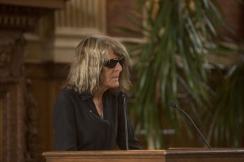 Joy Williams standing at a podium in black mourning clothes and wearing dark sunglasses while inside speaking at Gass' memorial service. 