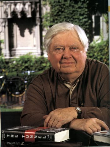 Photo of William H. Gass seated leaning at a table with his arms crossed in front of him and a copy of his book "The Tunnel."