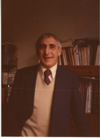 Waist-up photo of Raymond Federman. Federman is wearing a collared shirt and tie under a v-neck pullover with a suit jacket over the whole ensemble. Federman is smiling in the photo as he stands in a study. 