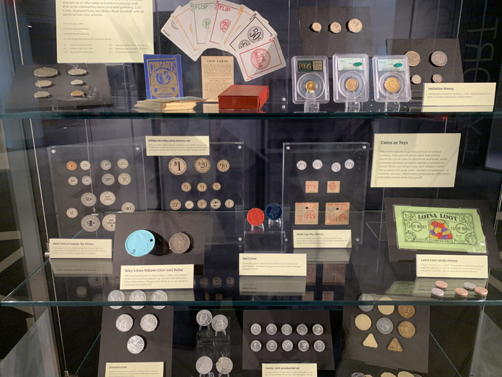 A closeup photo of one of the cases in the Worlds of Imagination exhibition in the showcases of the Newman Tower of Collections and Exploration. This case includes items from the Numismatics Collection and includes board game money.