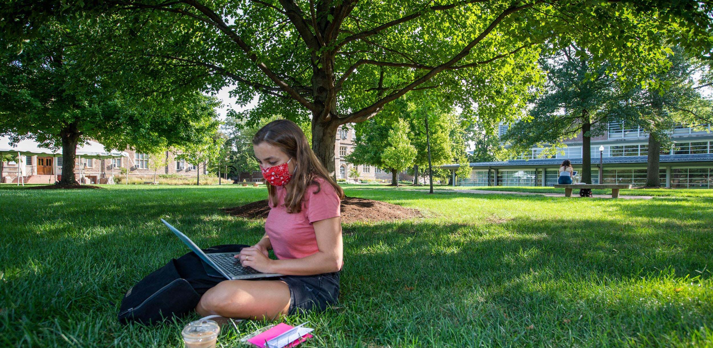 Image shows a student wearing a mask with a computer studying on a campus lawn.