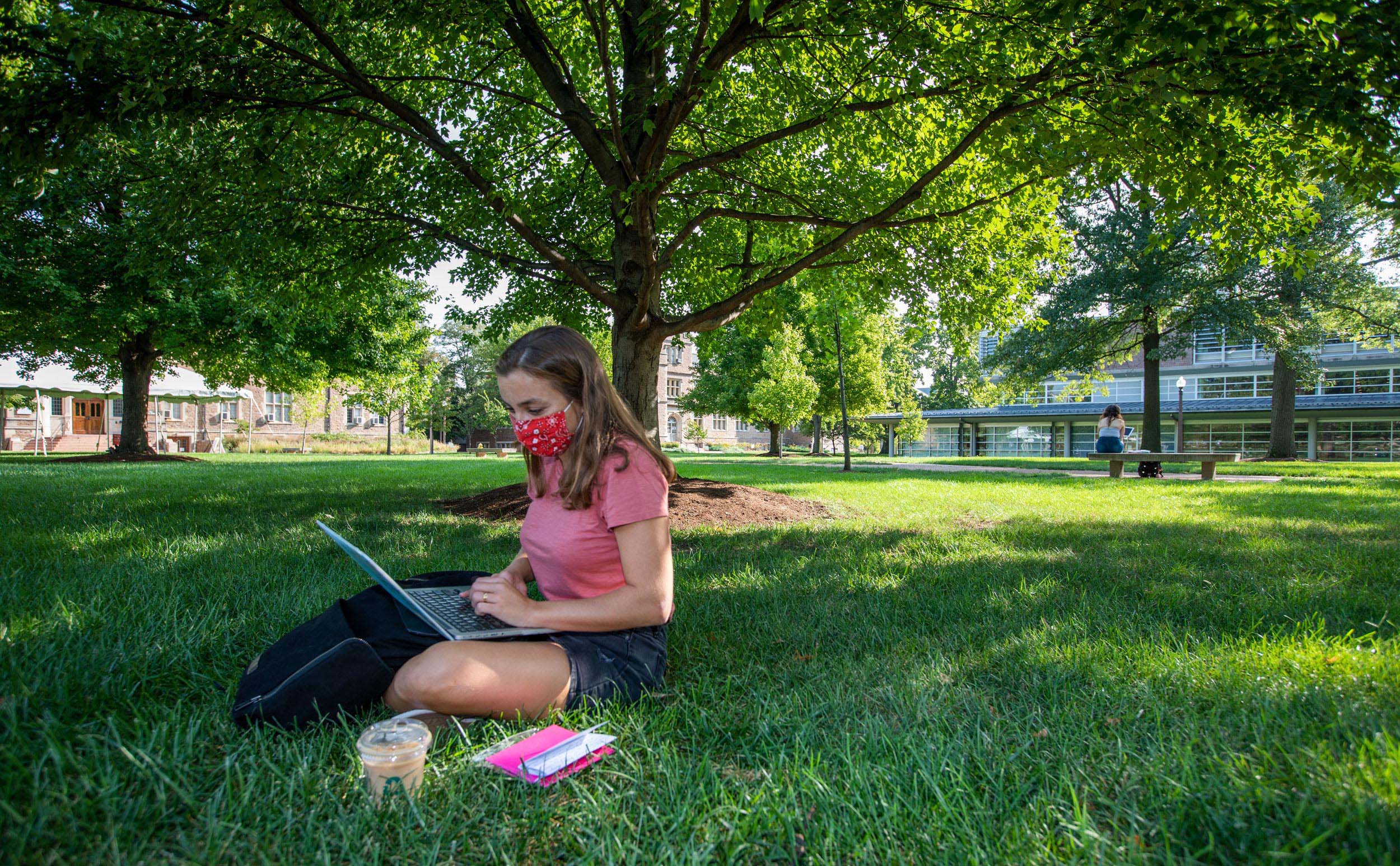 Image shows a student wearing a mask with a computer studying on a campus lawn.