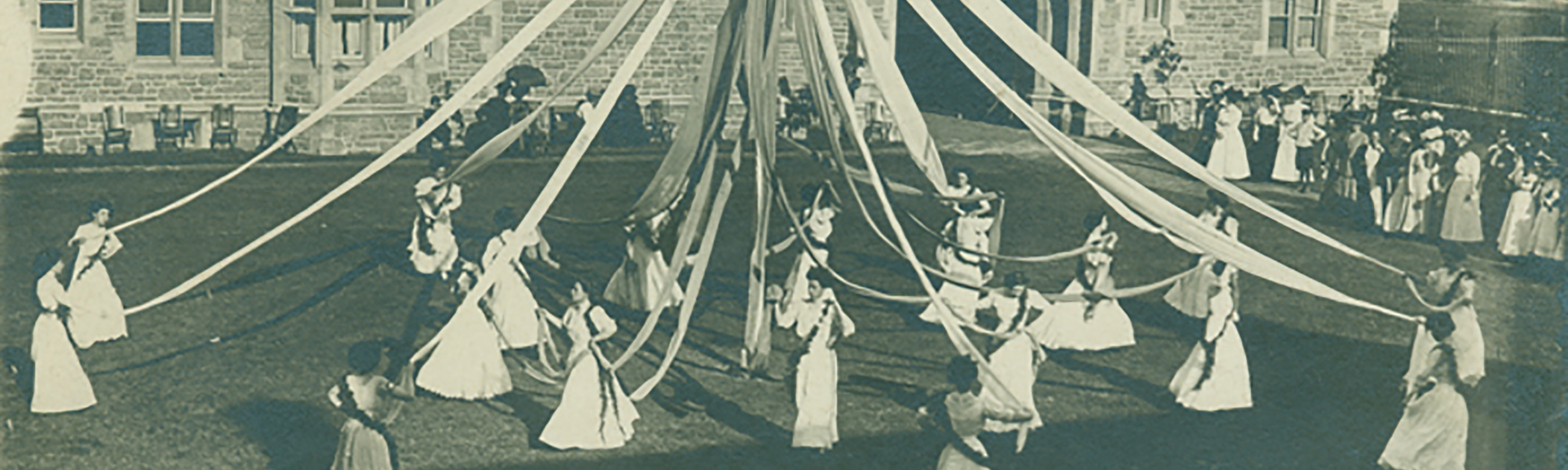 Header image shows a photo of female students dressed in white downs dancing a May Pole.