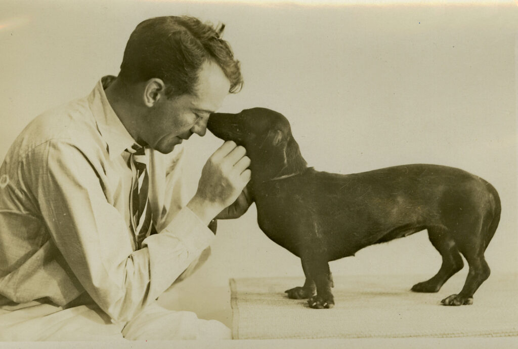 John Held Jr. is dressed in a button-down shirt with a tie and sits with his forehead touching his dog's nose. The dog is standing atop a table.