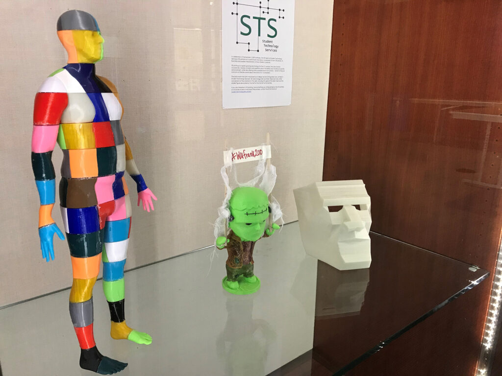 Three 3D printed works on display. One is a human figure printed in patch-work colors to represent Frankenstein's monster; another is a cartoonish depiction of a small, green version of Frankenstein's monster; and the last is the bust of Frankenstein's monster's head, depicted with sharp angles and harsh lines. 