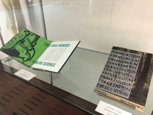 Two books in the "Frankenstein Experimental Book Design" submission by Gillian Fink. One book is open and displays artwork done on a green page; the other book is closed with the front cover displaying "FRANKENSTEIN" written repeatedly with a backdrop of a woods. 