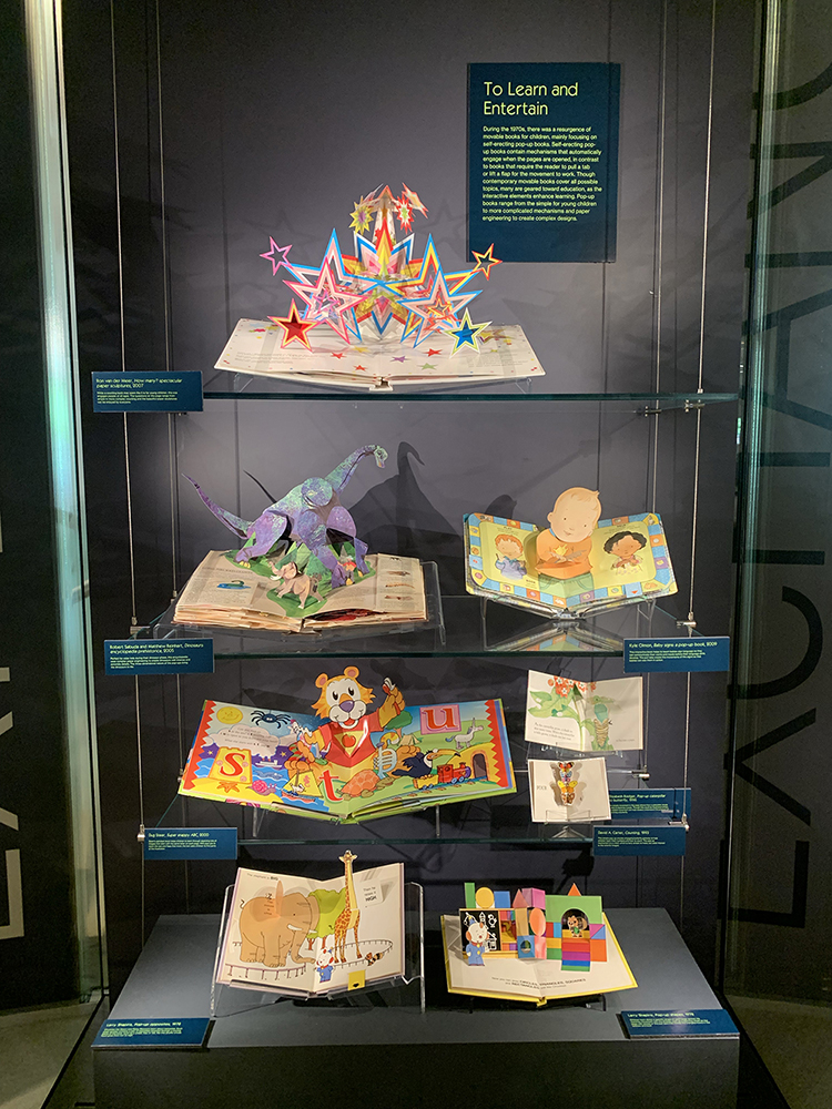 Display titled "To Learn and Entertain," part of the Moving Magic exhibition. The display features eight colorful, educational popup books of various sizes.