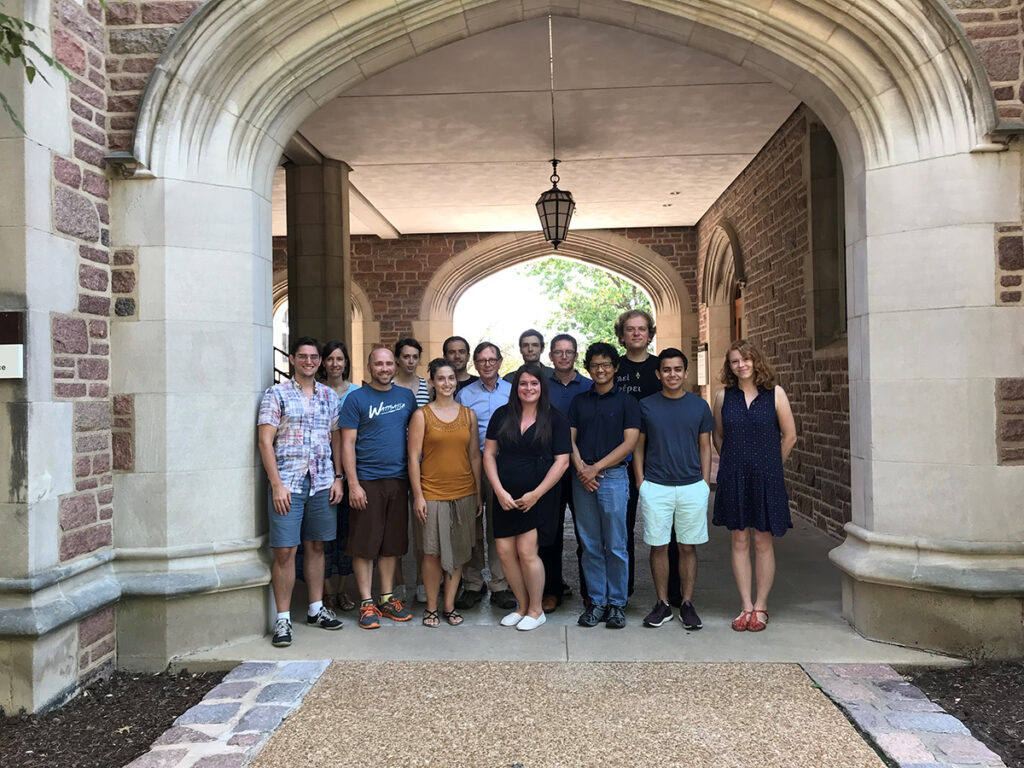 A group of fourteen stand under one of the arched walkways outside of the quad on the Washington University campus.