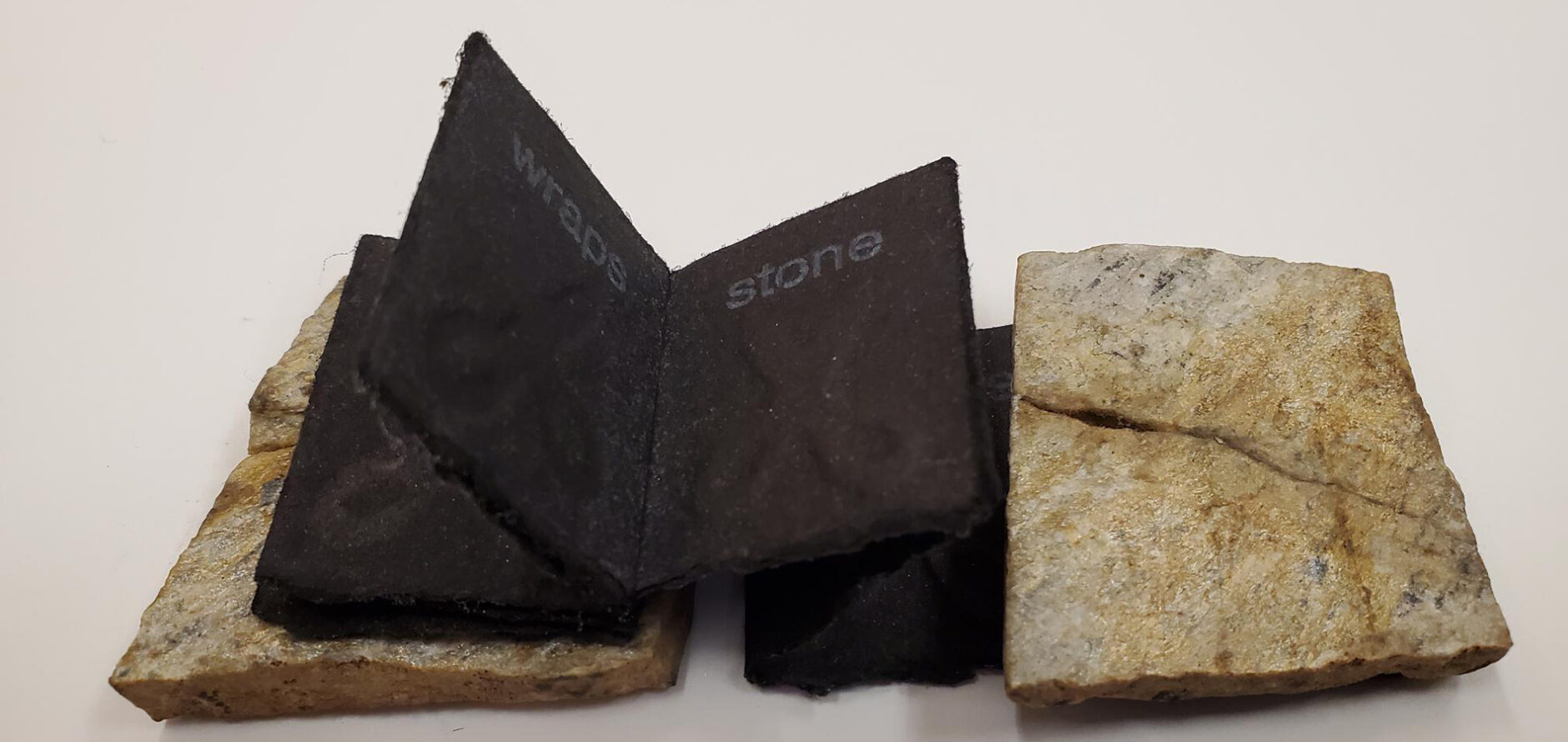 A miniature book with rocks as the covers and black, felt paper pages with impressions of scissors folded between the covers. Rock, paper, and scissors.