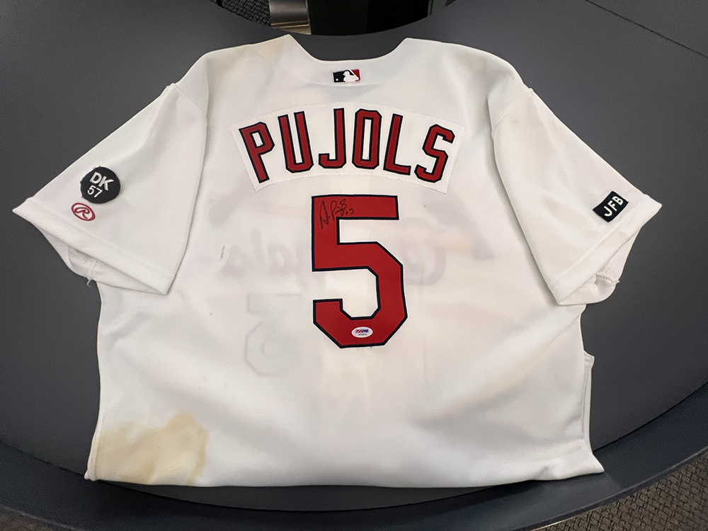 Signed game-worn Albert Pujols number 5 jersey with dirt stains on the bottom left hem. The jersey has a DK 57 circle emblem on one sleeve and JFB on the other. The jersey is in white.