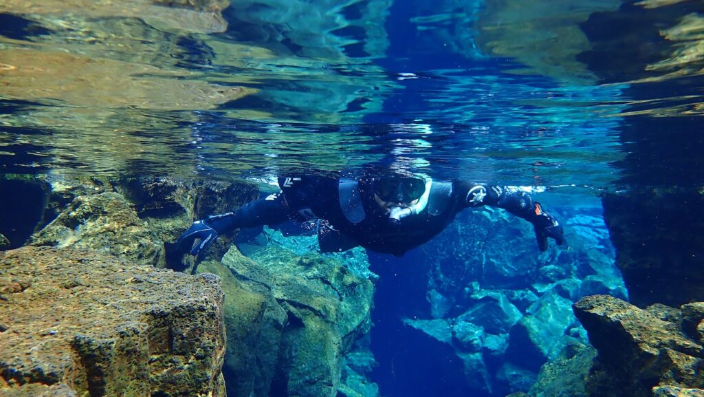 Person in a diving suit under blue lake water with rock formations on either side.
