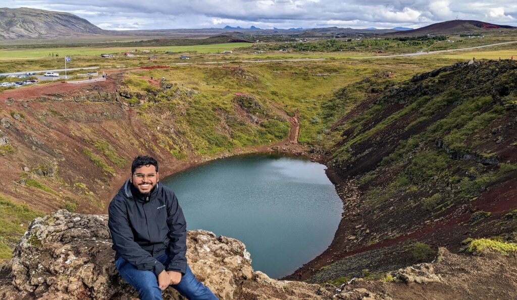 Person sitting on a rocky ledge overlooking a crater-shaped lake and greenery.