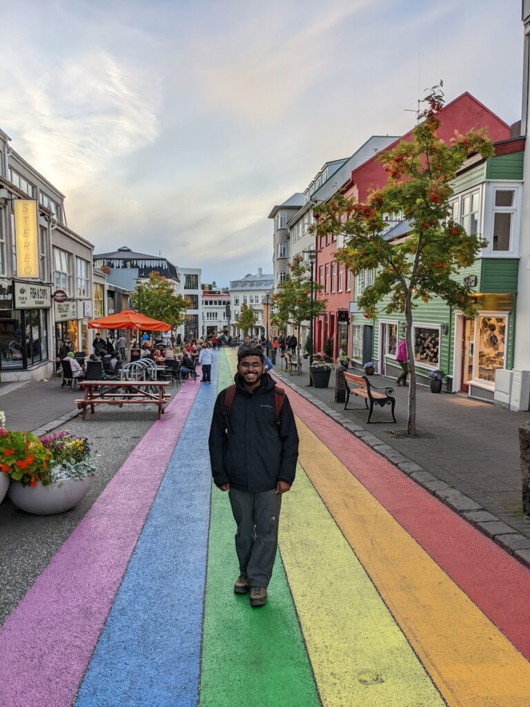 Person standing on a street painted in the colors of the rainbow with sidewalks and buildings on both sides.