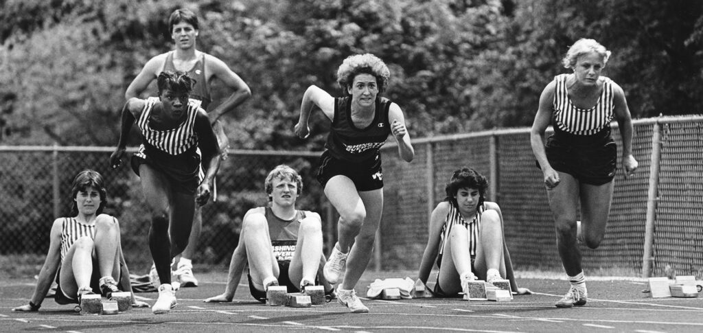 Three female track runners leaving the block as four of their teammates watch.