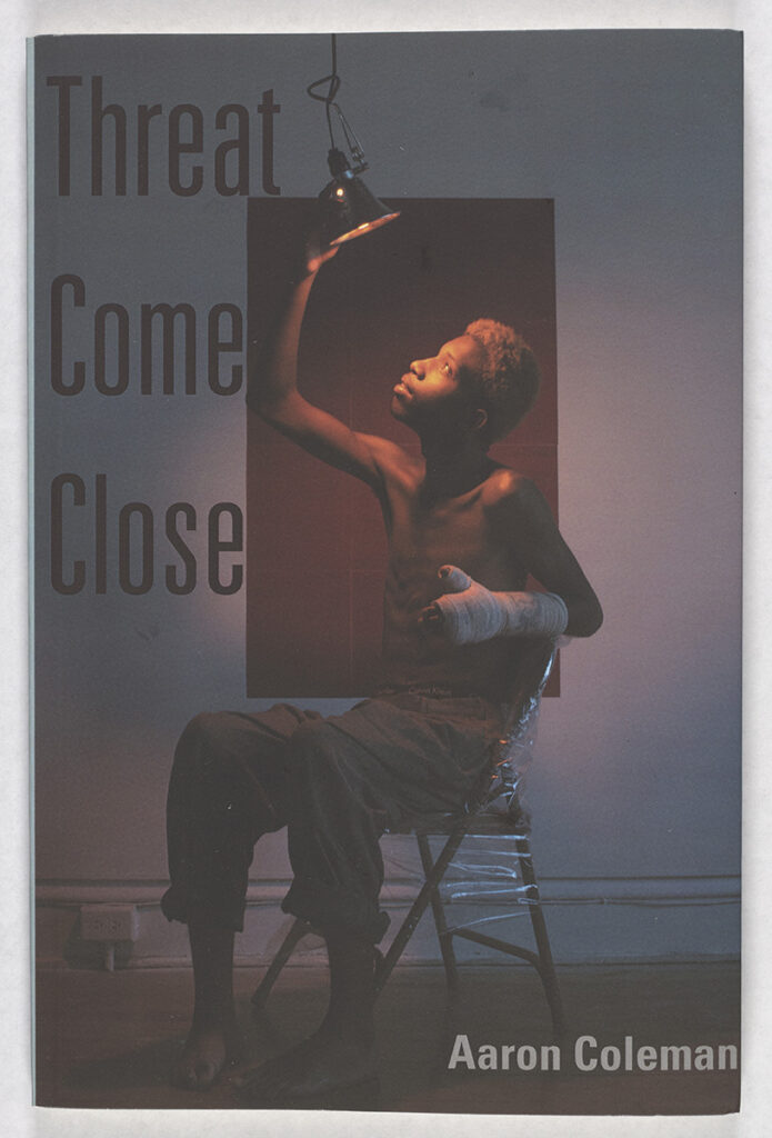 The covert art for Threat Come Close featuring a young Black man with a cast on his left arm seated in a metal folding chair reaching up to angle a heatlamp towards his upturned face.
