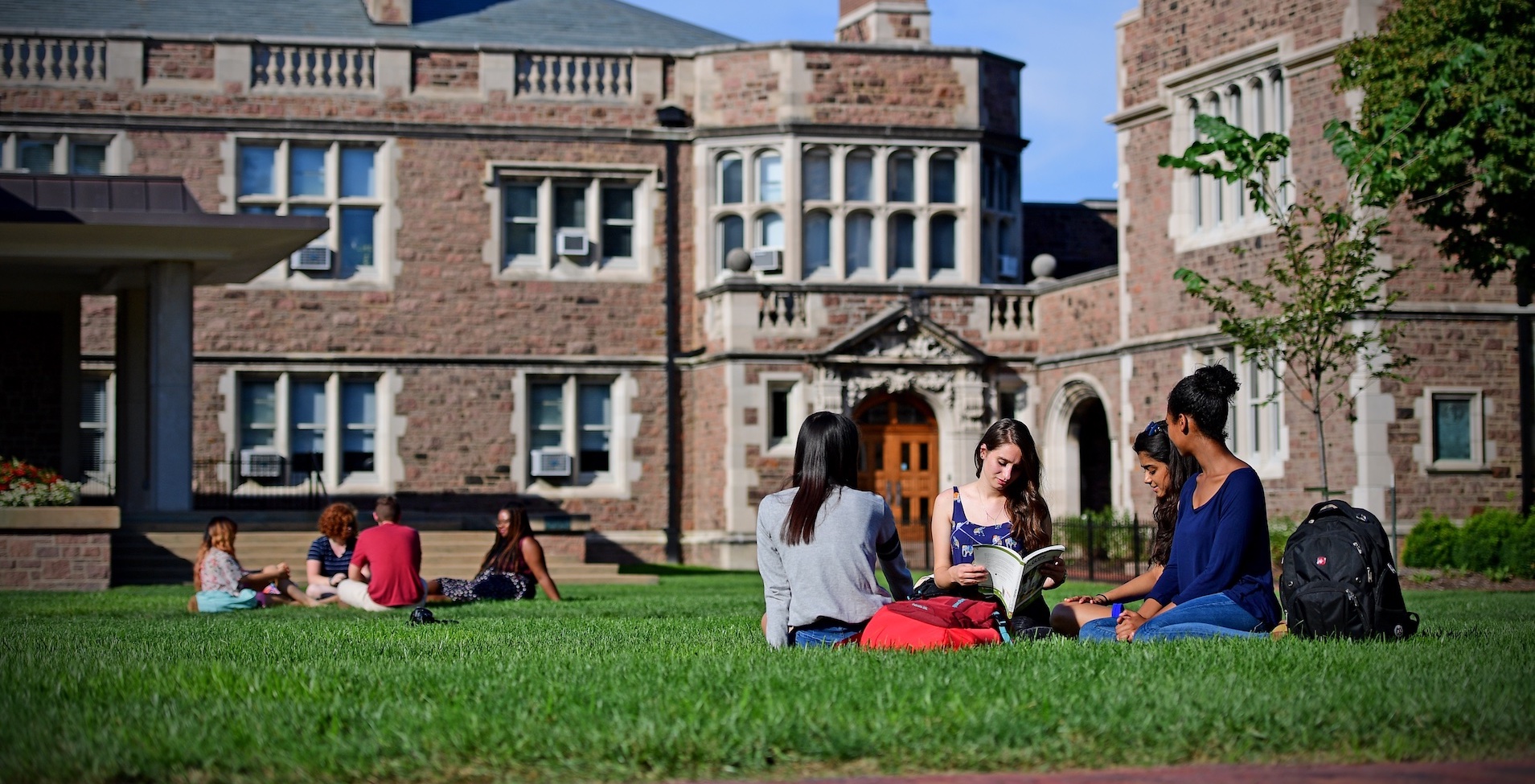 Two groups of studnets sitting and reading on the grass outside campus buildings