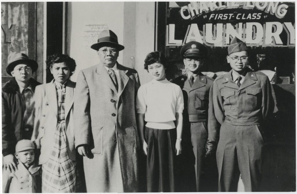 Black and whote photo of Asian Americans standing in front of a glass front store with the sign "First Class" Laundry