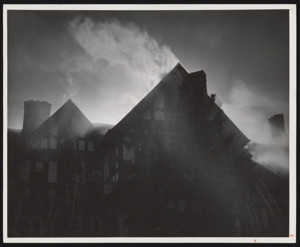 Black and white photo of a building with two triangular roofs with smoke rising out of the front portion of the building.
