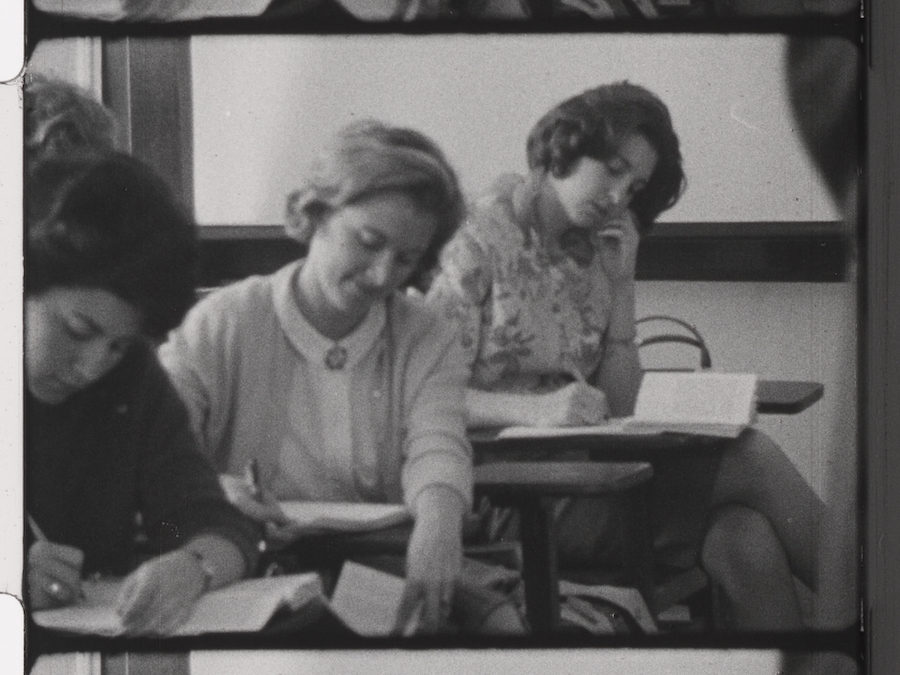 B&W photo of female student seated at classroom desks and writing.