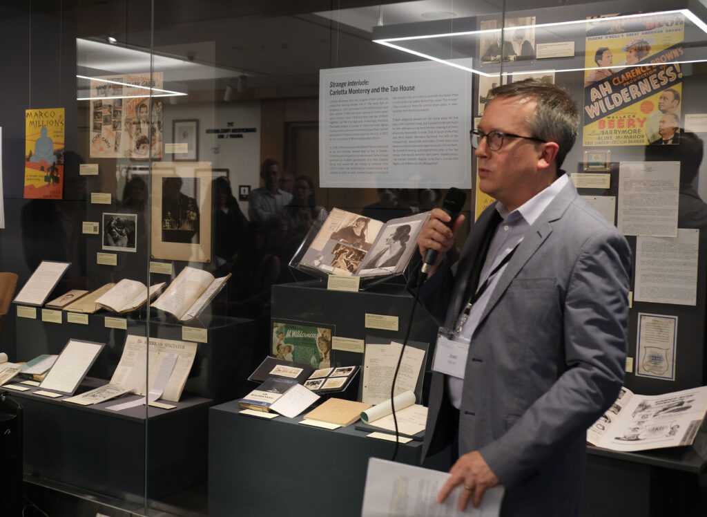 A man speaking into a mic in front of an exhibit displaying books and pictures.