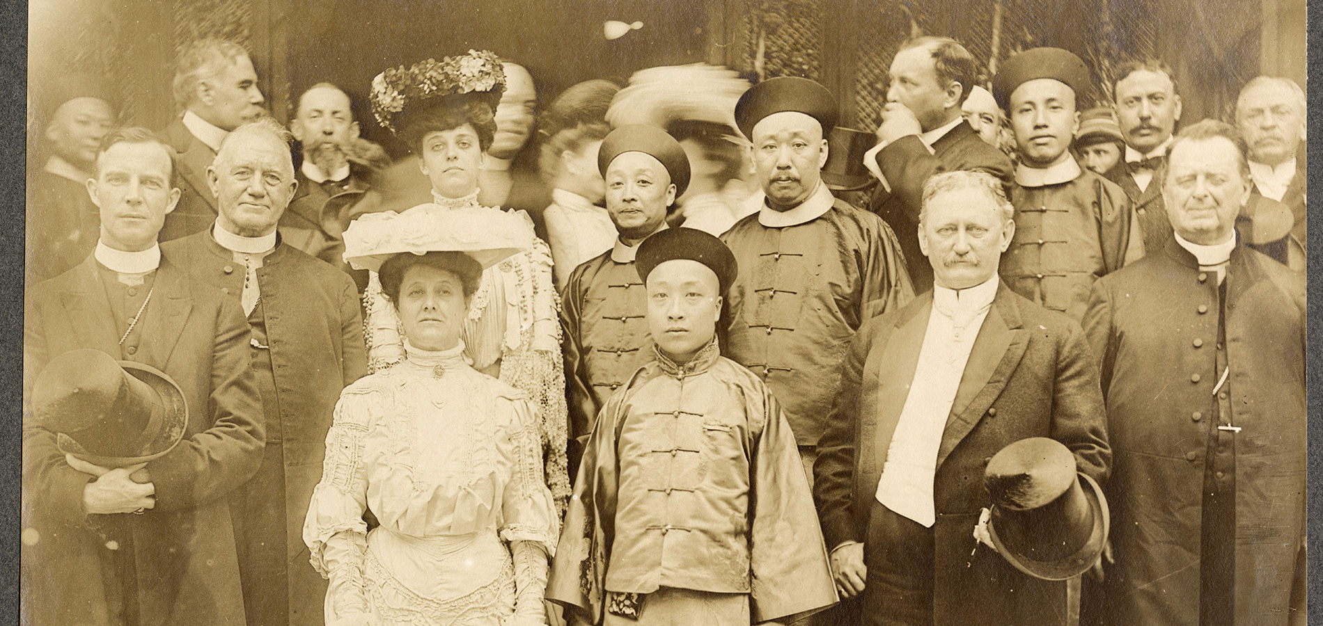 Four men of Chinese descent in traditional clothing standing for a photograph amidst a crowd of white men in dour suits and women in high-necked lace dresses with elaborate hats at the 1904 World's Fair.