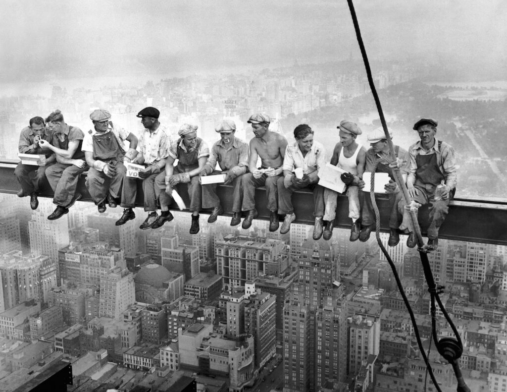 Men sitting on a beam high above the city skycrapers