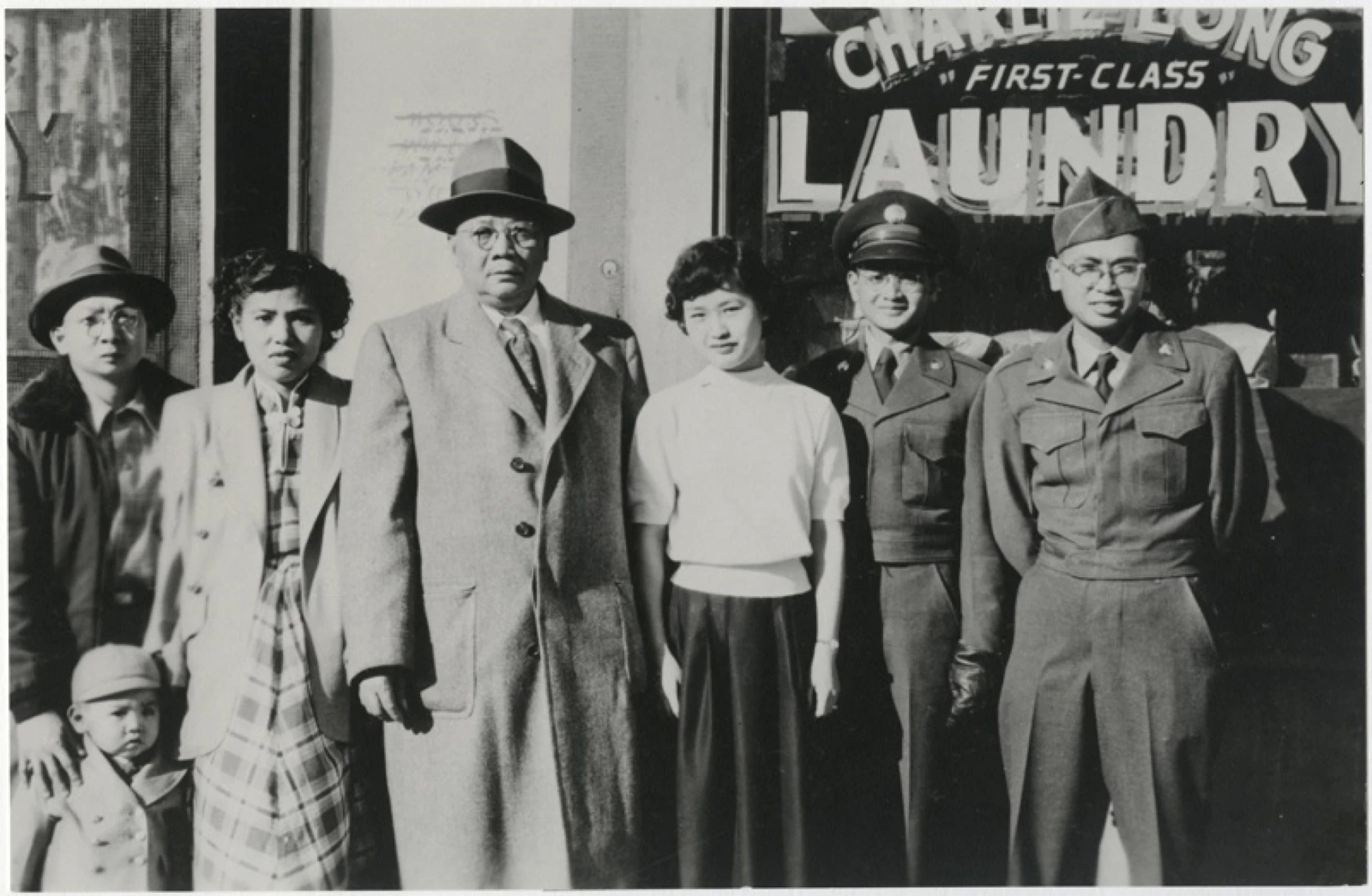 Four men, two women, and a child stand in front of a business. Two of the men are in uniform.